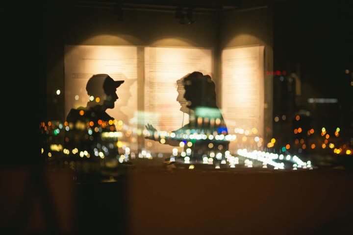 Due persone di profilo, in controluce, si parlano; sulle loro sagome si sovrappongono le luci sfocate di una città notturna e le pagine illuminate di un grande libro aperto