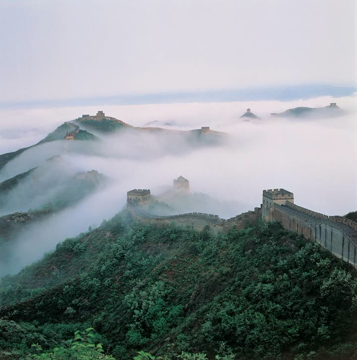 Un tratto di Grande Muraglia cinese corre sul crinale di colline boscose che emergono da un mare di nebbia; in lontananza altre torri affiorano appena dalle nuvole
